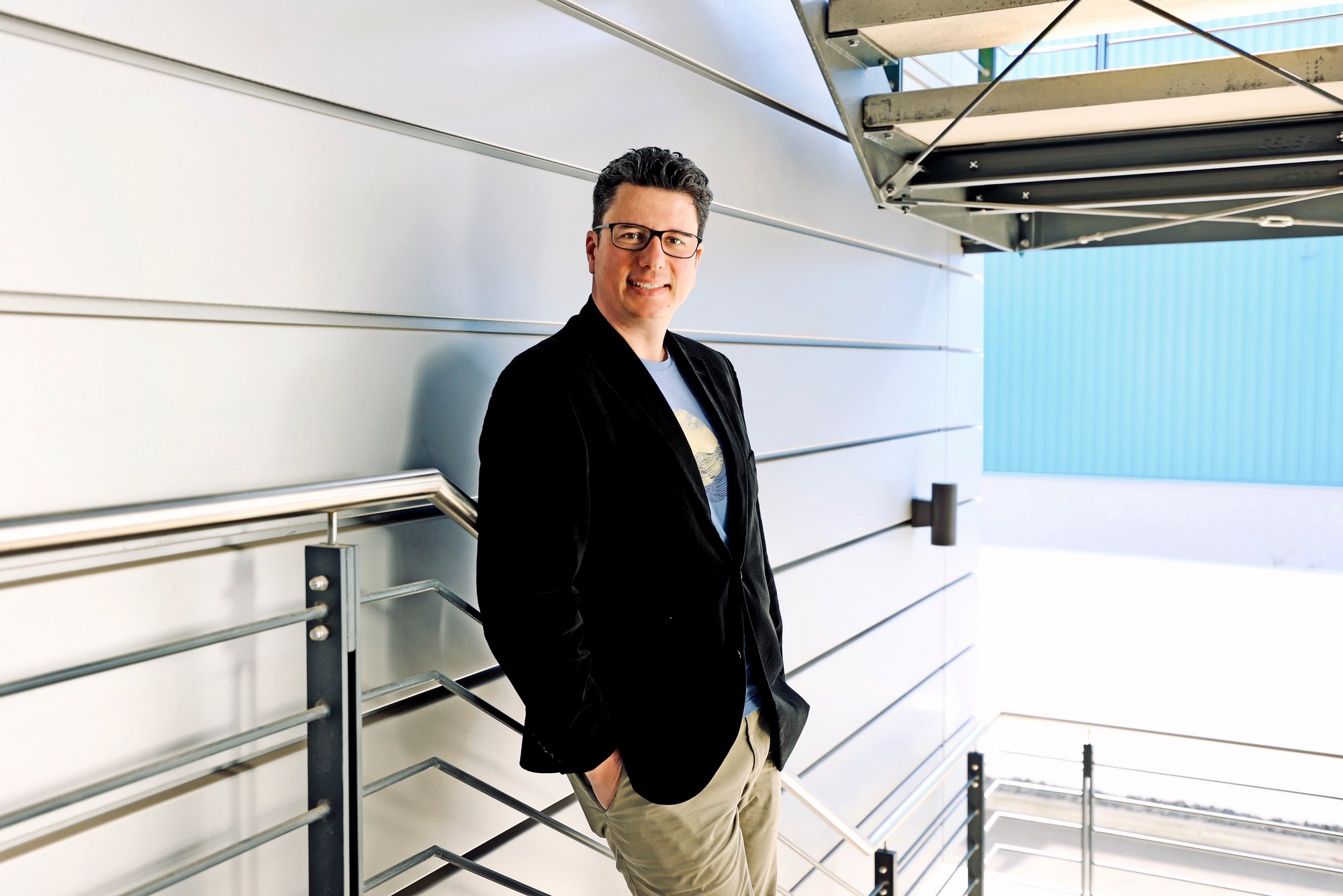 Porträtfoto eines Mitarbeiters, der entspannt an einem Geländer lehnt. Er trägt legere Businesskleidung und blickt freundlich in die Kamera. Die Umgebung wirkt modern.