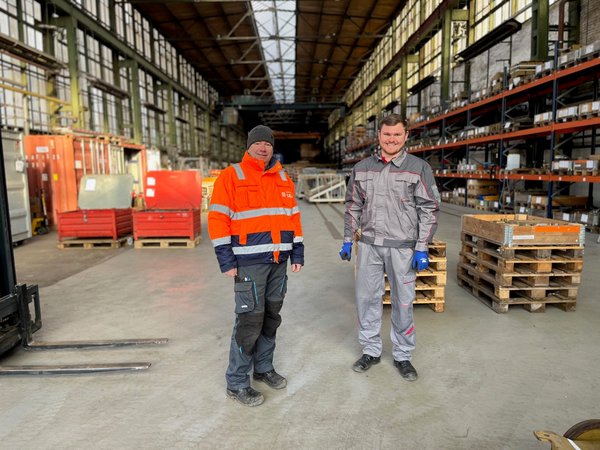 Zwei Lagerarbeiter vor Regalen und Paletten, einer in Orange, der andere in Grau gekleidet