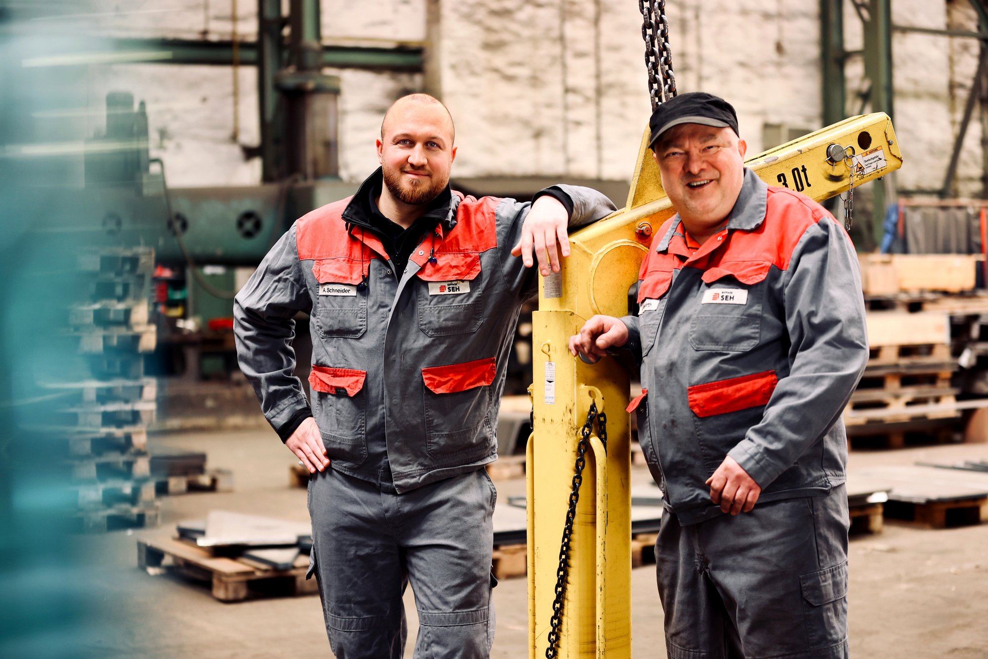 Zwei Männer in Arbeitskleidung, freundlich lächelnd in einer Industrie- oder Baustellenumgebung