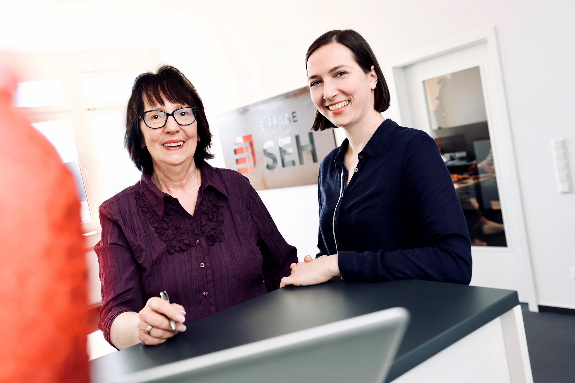 Zwei Frauen vor einem Tresen, eine mit Brille und lila Bluse, die andere mit dunkelblauer Bluse. Beide lächeln.