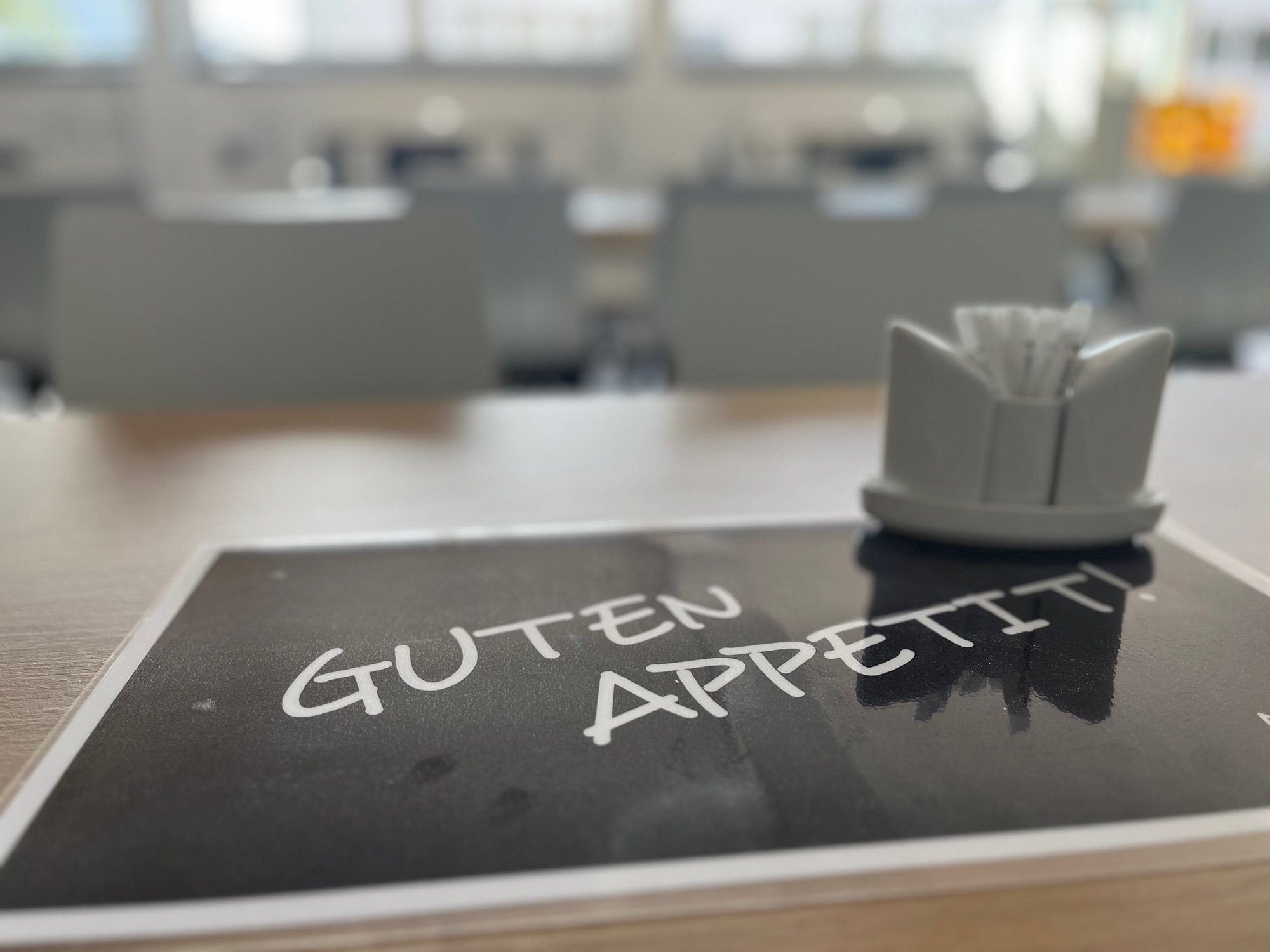 Tischset oder Zettel mit „Guten Appetit“-Schriftzug, Kantinenumgebung im Hintergrund