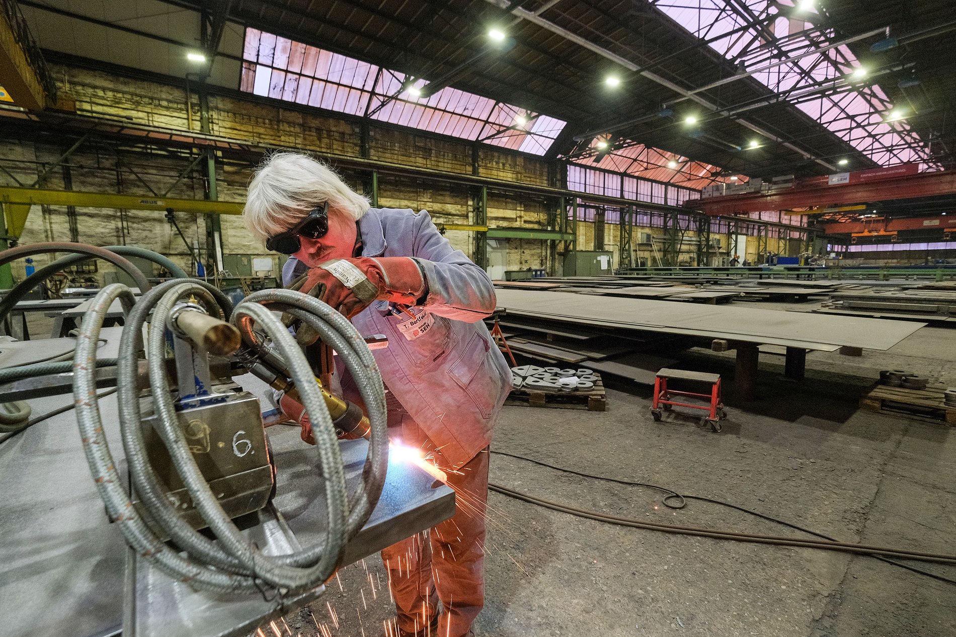 Mitarbeiter in Arbeitskleidung mit Schutzbrille und Handschuhen in einer Produktionshalle, bedient ein Gerät mit Schlauch, aus dem Funken austreten